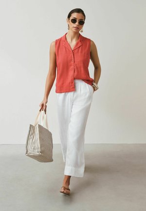 Woman wearing round sunglasses, sleeveless rust-colored button shirt, white wide-leg pants, carrying striped tote bag, walking forward indoors.