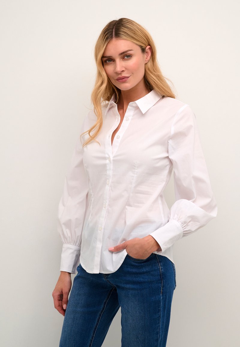 Vit skjorta med klassisk krage och puffärmar, tillverkad av ett slätt tyg. Buren med blå denimjeans, händerna på höfterna.