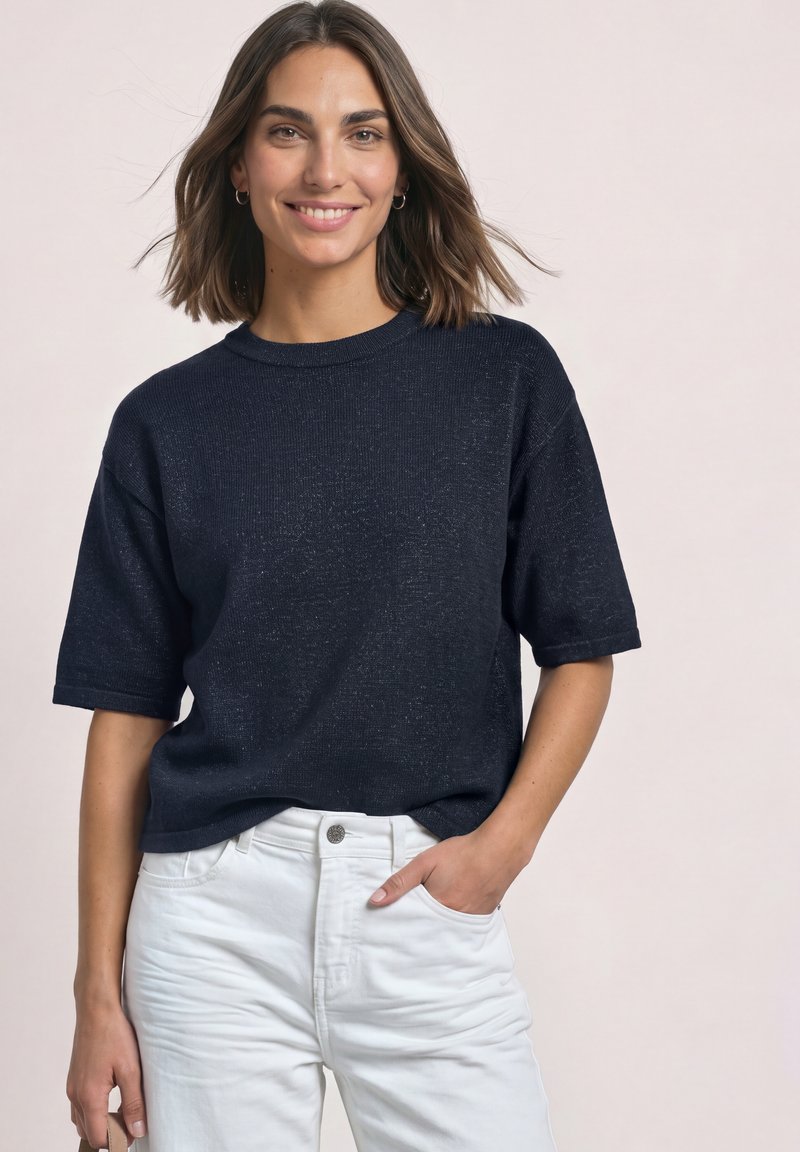 Femme souriante aux cheveux bruns mi-longs, portant une chemise bleu marine à manches courtes et un pantalon blanc, une main dans la poche.