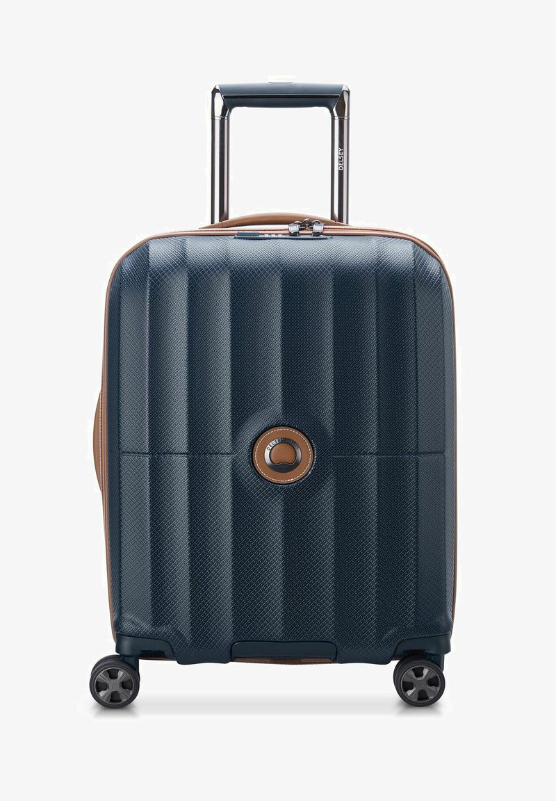 Valigia rigida di colore blu scuro con superficie testurizzata, dettagli in beige, manico retrattile e quattro ruote per la mobilità. Dettaglio con logo circolare.