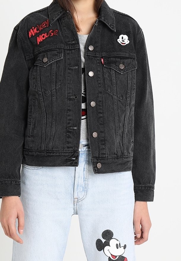 Veste en jean noire avec texte rouge "Mickey Mouse" et petite écusson Mickey, portée avec un jean bleu clair arborant une grande impression de Mickey Mouse.