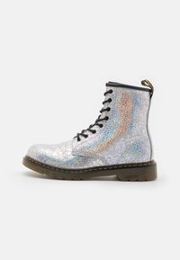 Srebrne buty wojskowe z pękającym holograficznym wykończeniem, czarne sznurowadła, gumowa podeszwa i żółte przeszycia. Posiadają pętelkę na pięcie.