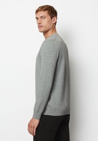 Mann mit kurzen blonden Haaren, der einen hellgrauen Langarmpullover und schwarze Hosen trägt, seitlich vor einem weißen Hintergrund stehend.