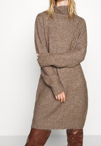 Robe-pull tricotée marron avec un col roulé haut, des manches longues et une coupe décontractée, présentant une texture douce et des poignets côtelés.