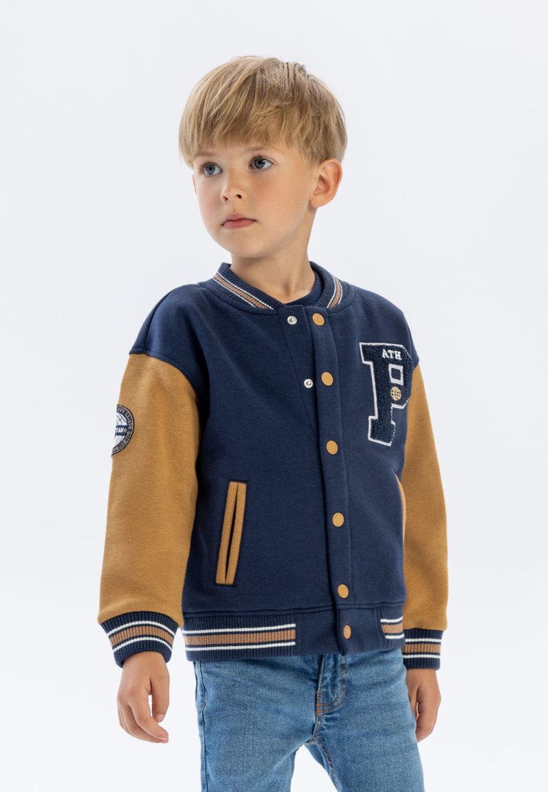 Blaues und sandfarbiges Varsity-Jackett mit Druckknöpfen, gestreiften Bündchen, gesticktem Patch und Seitentaschen. Getragen über blauen Jeans.