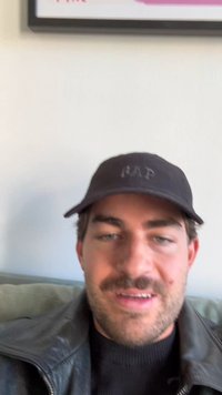 Pessoa usando um boné preto com o logotipo "GAP", estilizada com uma jaqueta de couro, sentada num sofá verde contra uma parede clara.