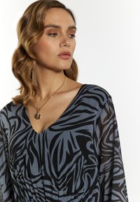 Pitkähihainen sininen mekko mustalla abstraktilla kuviolla, jossa on V-pääntie. Saatavilla oleva kangas on sileä ja miellyttävä. Korostuksena kultaketju.