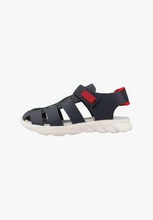 Navy blue strappy sandals with a textured rubber upper, white cushioned sole, and red branded accent. Adjustable velcro strap for fit.