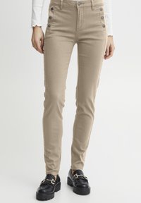 Pantalon beige taille haute à la coupe ajustée, avec des accents de boutons sur les côtés et une texture lisse. Porté avec des mocassins à plateforme noirs.