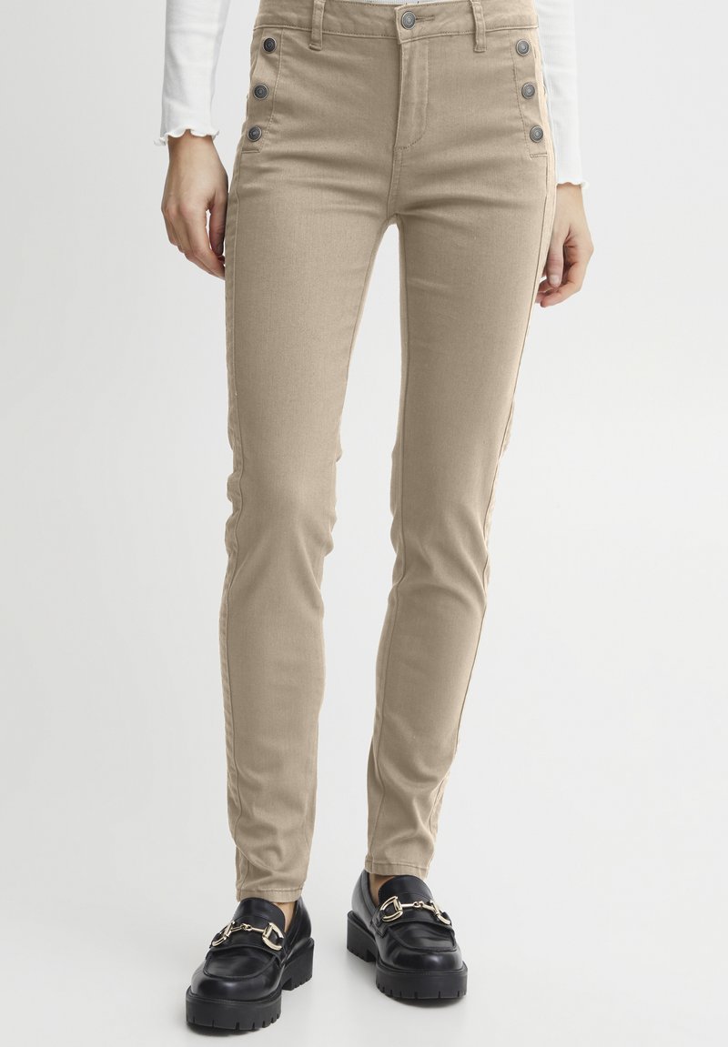 Pantalon beige taille haute à la coupe ajustée, avec des accents de boutons sur les côtés et une texture lisse. Porté avec des mocassins à plateforme noirs.