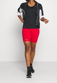 Giacca sportiva nera con accenti bianchi, canotta nera e pantaloni corti rossi aderenti. Presenta loghi Nike sui capi d'abbigliamento e un design aerodinamico.
