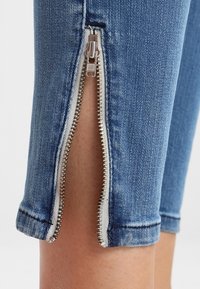 Blå denimjeans med en silverfärgad metalldragkedja vid fållen, med en närbild av den delvis öppna dragkedjan och den sydda kanten.