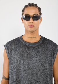 Black oval sunglasses with a sleek frame, worn with a gray sleeveless t-shirt featuring a textured fabric. Notable tattoo visible on the neck.