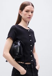 Μαύρο τοπ με κοντά μανίκια και κυματιστές άκρες, με στρογγυλή λαιμόκοψη και μαργαριταρένια κουμπιά μπροστά. Συνοδεύεται από μαύρη τσάντα με πιέτες.