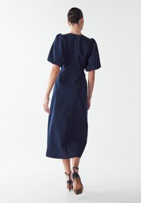 Mørkeblå midi-kjole med pufærmer, tætsiddende talje og lynlås i ryggen. Stylet med sorte højhælede sko og minimale smykker.