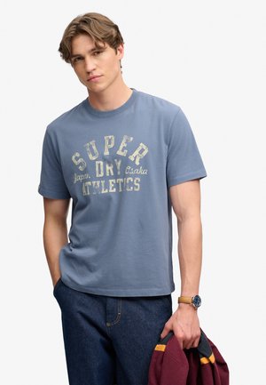 Ung mand med kort bølget hår iført en blå "Super Dry Athletics" t-shirt, mørke jeans, et armbåndsur og holder en mørkerød jakke.