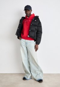 Black puffer jacket, red hoodie with logo, light blue wide-leg jeans with frayed hem, black shoes, and a navy cap.