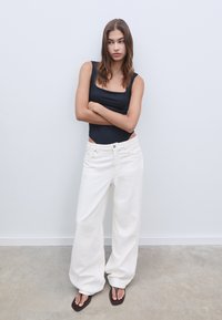Svart ärmlös bodysuit i kombination med lösa, hög midja vita jeans i denim. Modellen bär bruna flip-flops, med avslappnad passform och mjuk textur.