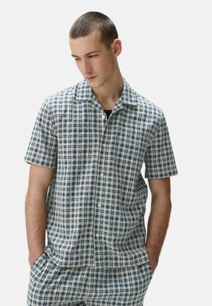 Mężczyzna w niebiesko-białej koszuli na guziki w kratę z krótkim rękawem i pasujących spodniach, ręce w kieszeniach, patrzący w dół i w lewo.