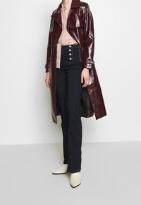 Trench-coat bordeaux brillant avec un col à revers, associé à un jean évasé fonc é orné de boutons décoratifs et des bottines crème.