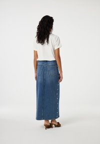 Blauwe denim maxi-rok met een rechte pasvorm, twee achterzakken en knopen aan de zijkant. Gecombineerd met een wit topje met korte mouwen en stevige hakken met patroon.