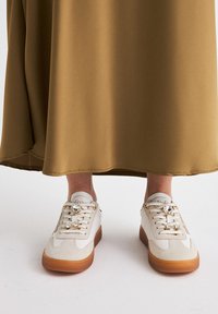 Zapatillas blancas y beige con un acabado texturizado, que cuentan con piedras preciosas decorativas en los cordones y una suela de goma marrón. Llevadas con una falda larga marrón.