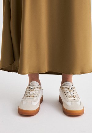 Zapatillas blancas y beige con un acabado texturizado, que cuentan con piedras preciosas decorativas en los cordones y una suela de goma marrón. Llevadas con una falda larga marrón.
