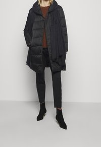 Abrigo negro acolchado con capucha, diseño acolchado, combinado con un suéter marrón, pantalones negros y botines negros, con una bufanda texturizada.