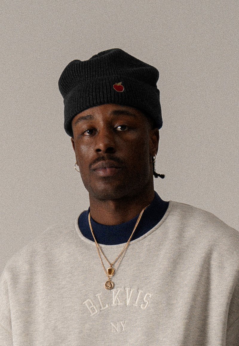 Beanie di maglia nera con dettaglio di ricamo a forma di ciliegia. Felpa grigia con testo "BLKVIS NY", indossata sopra un colletto blu scuro. Collane d'oro visibili.
