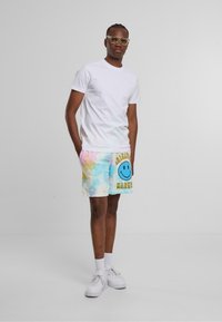 Hvid t-shirt kombineret med multifarvede tie-dye shorts med et blåt smileyface grafik og gult tekst, suppleret med hvide sneakers.