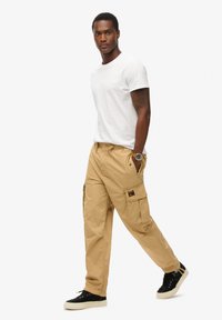 Pantalons cargo beiges avec ceinture élastique, plusieurs poches et un patch logo. Associés à un t-shirt blanc ajusté et des baskets noires.