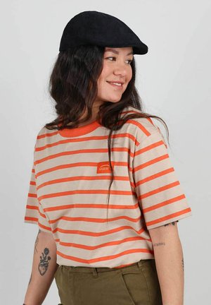 T-shirt beige à rayures avec des rayures orange et un petit patch logo. Col ras du cou, manches courtes et coupe décontractée. Porté avec une casquette noire.