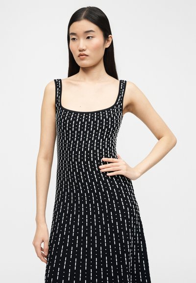 Ασιάτισσα με μακριά μαύρα μαλλιά, ντυμένη με μαύρο αμάνικο φόρεμα με λευκές κάθετες διακεκομμένες γραμμές, στέκεται με το ένα χέρι στη μέση.