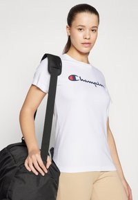 Bijela pamučna T-majica s "Champion" logom u tamnoplavoj i crvenoj boji. Tamnosmeđa torba s teksturiranim remenom. Siva pozadina.
