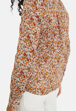 Mujer con blusa de manga larga con un estampado floral denso en tonos naranja, rojo, rosa y verde, combinada con pantalones blancos, sobre un fondo blanco.