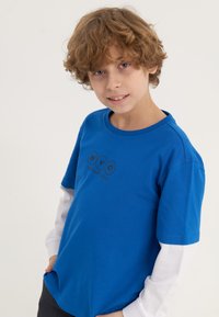 Blue cotton t-shirt with black “NYC New Youth Culture” logo, crew neckline, short sleeves, layered with a white long-sleeve shirt underneath.