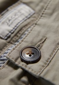 Lähetä beigen värinen kangastakki lähikuvassa, jossa on kiiltävä tumma nappi, ompelutiedot ja merkkilappu yläreunassa.