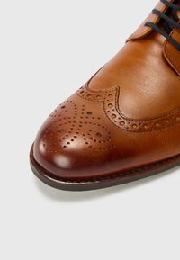 Zapato de vestir de cuero marrón con punta redondeada, perforaciones de brogue detalladas, textura suave y cordones negros resistentes. Diseño clásico.