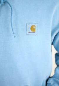 Light blue hoodie made of soft fabric, featuring a square white patch with a yellow logo on the front. Includes a drawstring hood.