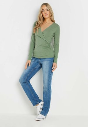 Langsleeve groene wikkeltop met plooidetails, gecombineerd met recht-toe recht-aan blauwe jeans en witte sneakers met zwarte strepen.