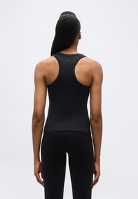 Γυναίκα που στέκεται με τ ην πλάτη, φοράει μαύρο αμάνικο τοπ με αθλητική πλάτη και μαύρο κολάν, μπροστά σε απλό ανοιχτό φόντο.