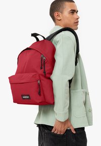 Mochila roja con textura suave, bolsillo frontal y correas negras ajustables. Etiqueta de la marca visible en la parte frontal. Llevada por una persona con una camiseta verde.