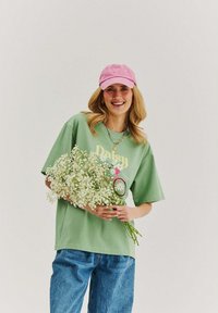 Young woman smiling, wearing green graphic t-shirt, pink cap, blue jeans, holding a large bouquet of white baby's breath flowers.