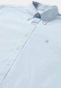 Světle modrá košile s knoflíky vyrobená z látky s hladkou texturou, s límečkem, béžovými knoflíky a vyšitým logem na hrudi.