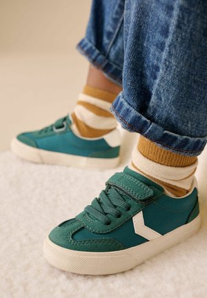 Žalios ir baltos sportbačiai turi tekstūruotą audinio viršutinę dalį, raištelius ir guminį pado sluoksnį, derantį su rudomis ir kreminėmis dryžuotomis kojinėmis.