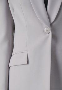 Blazer gris claro con tejido suave y elástico, cierre de un solo botón, solapas con muesca y un pequeño detalle de bolsillo en la parte delantera.