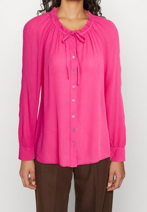 Une femme porte un chemisier rose vif à manches longues avec des boutons sur le devant et un col noué, associé à un pantalon marron, sur un fond uni.