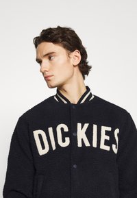 Giacca in fleece blu navy con lettere a contrasto in crema. Chiusura con bottoni a pressione, colletto a coste e texture morbida. Adatta per un abbigliamento casual.