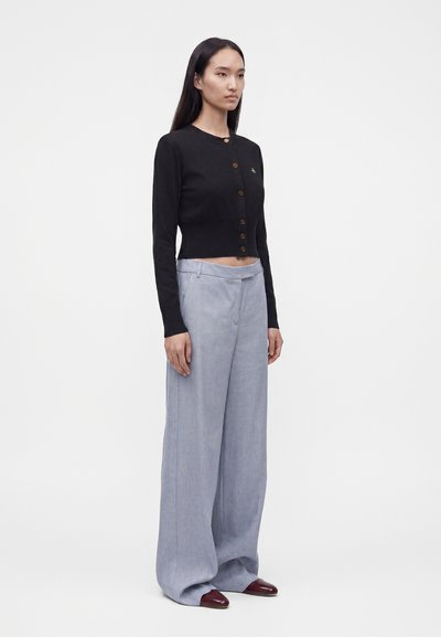 Γυναίκα που φοράει μαύρη κουμπωτή κοντή ζακέτα, ανοιχτό γκρι φαρδιά παντελόνια και σκουρόκόκκινα μυτερά παπούτσια, στέκεται μπροστά σε απλό φόντο.