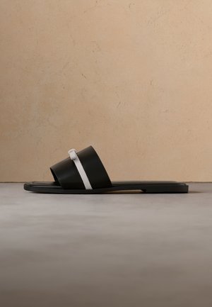 Sandale plate noire à enfiler avec une large lanière supérieure comportant une bande décorative blanche et une petite boucle, sur fond beige.